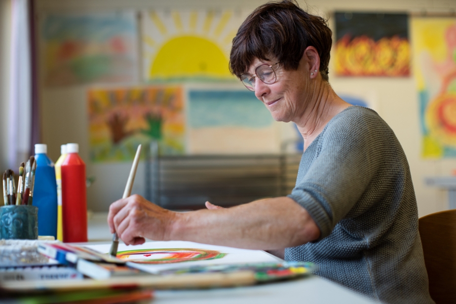 Eine etwas ältere Frau mit kurzen dunklen Haaren malt in der Therapiestunde mit einem Pinsel ein buntes Bild mit Wasserfarben.